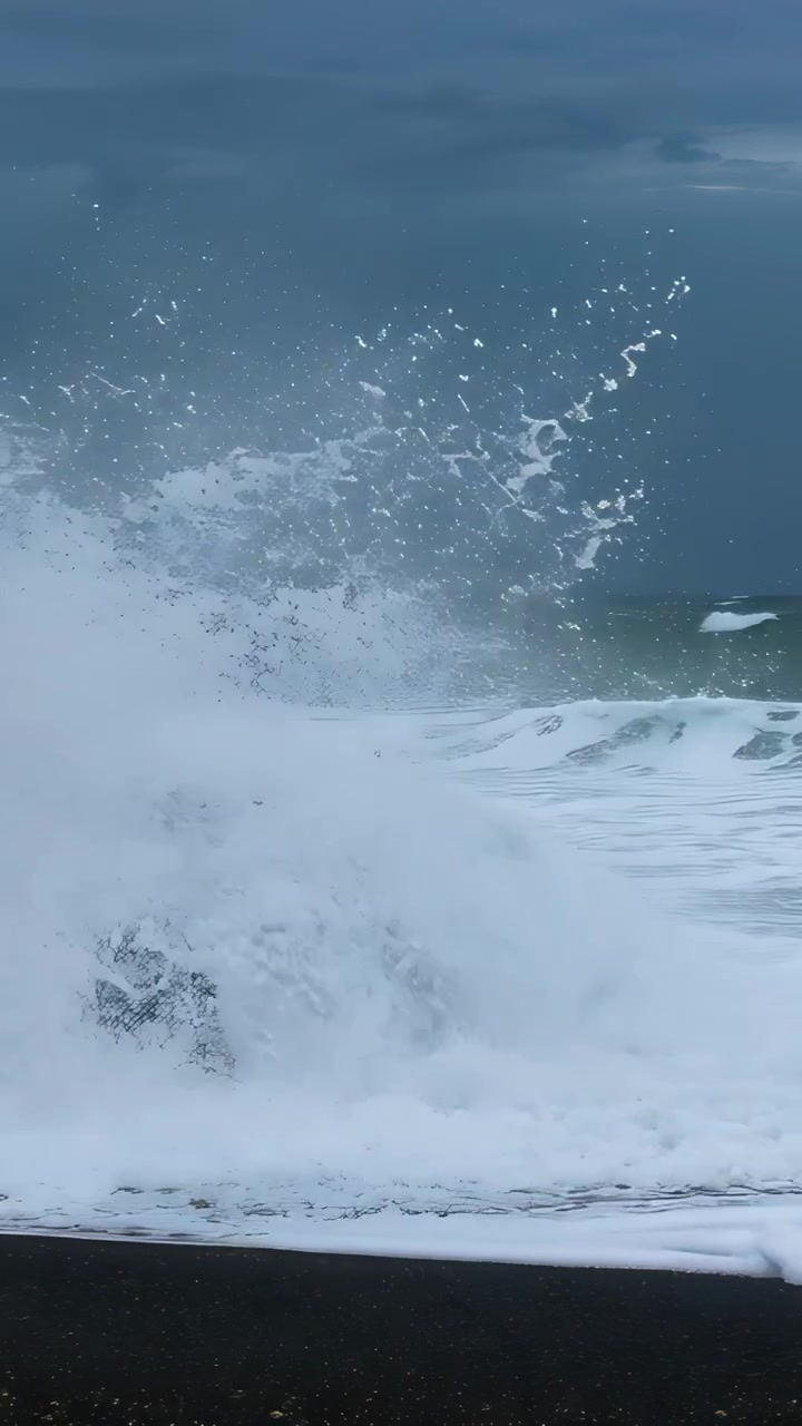 Vertical video: Approaching swell striking centered dark rock on black sand, sending spray skyward