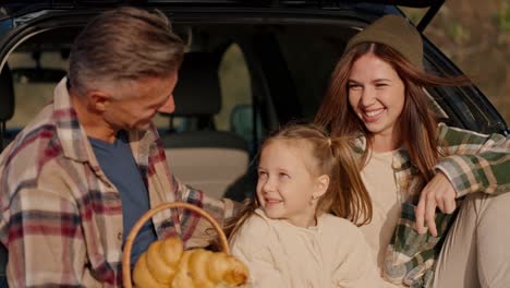 Happy-brunette-girl-in-a-hat-in-a-green-checkered-shirt-communicates-with-her-husband,-a-middle-aged-man-in-a-checkered-shirt,-and-her-little-daughter,-a-blonde-girl,-during-their-picnic-outside-the-city-near-the-car-in-the-summer