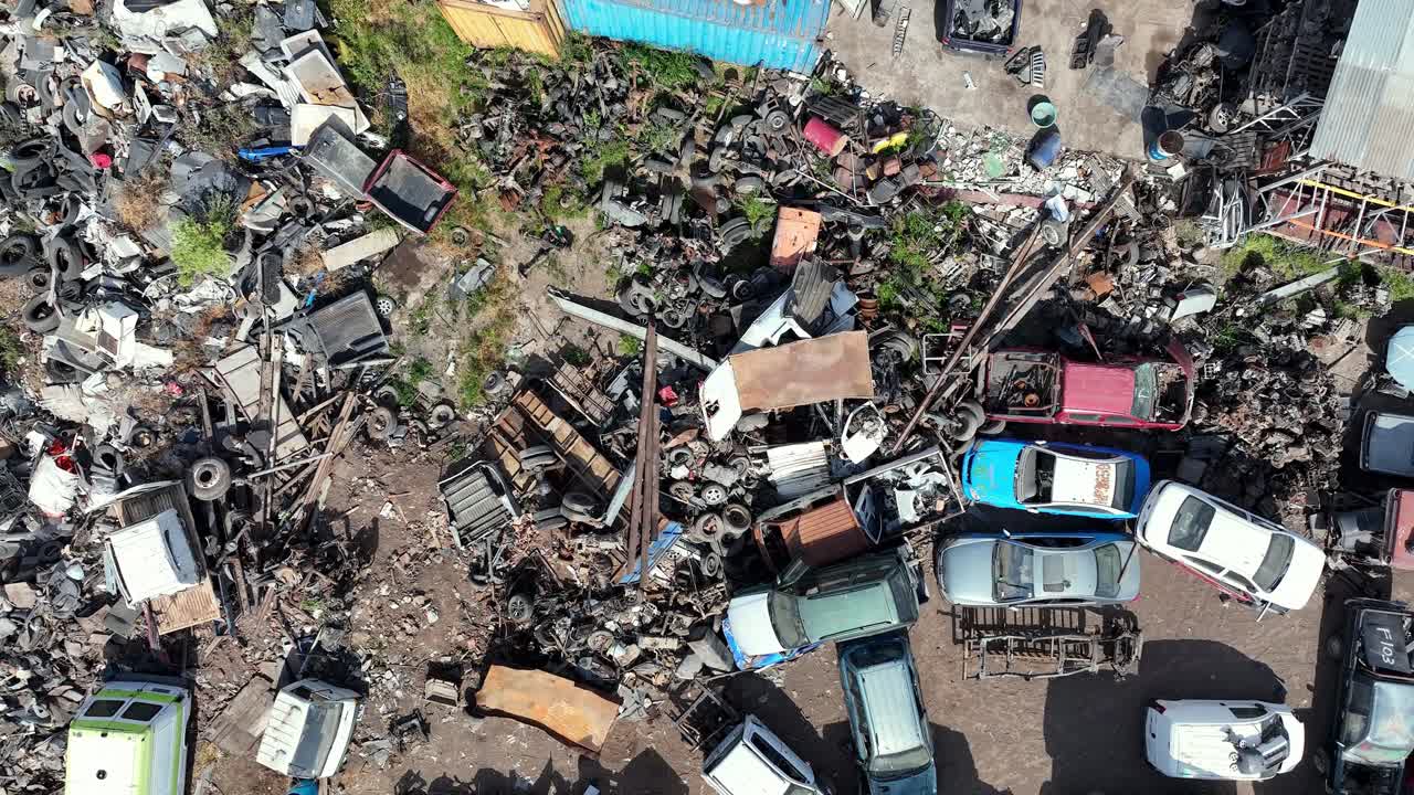 Forward drone aerial of chaotic junkyard with scrapped cars, trucks, auto parts, and metal debris piles in industrial salvage yard