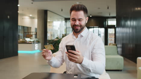 happy businessman using credit card and smartphone for online shopping payments in office lobby