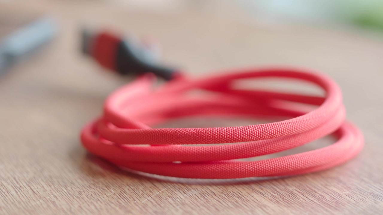 Hånd samler et rødt flettet USB-kabel op fra et træskrivebord