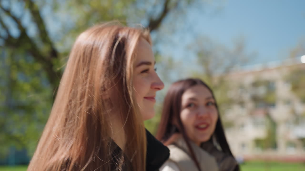 두 명의 여성, 백인과 아시아인이 평화로운 공원에서 하늘을 바라보고 있어요. 희망에 찬 표정과 부드러운 햇살, 잎이 무성한 나뭇가지와 멀리 보이는 건물들, 미래를 계획하고 공유된 꿈을 꾸는 사색적인 분위기예요.