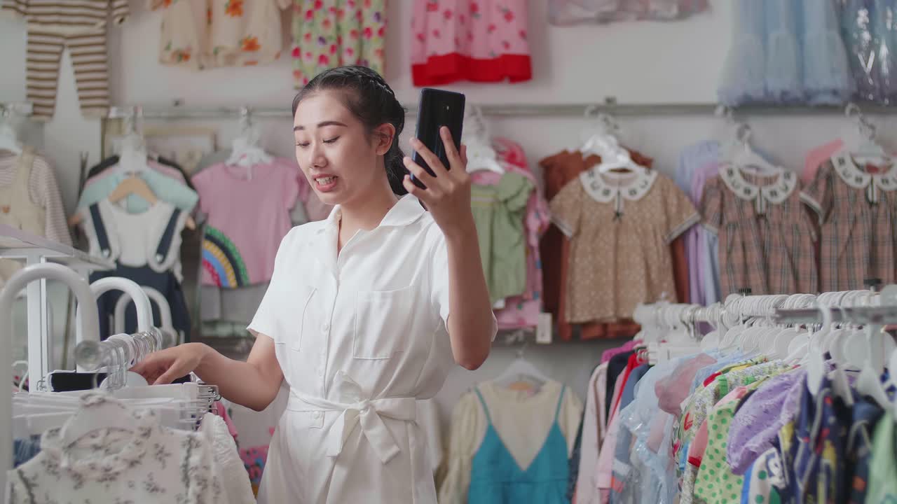 아시아 여성은 손을 흔들고 부티크에서 스마트폰으로 어린이 옷을 판매하는 스트림을 살고 있습니다.