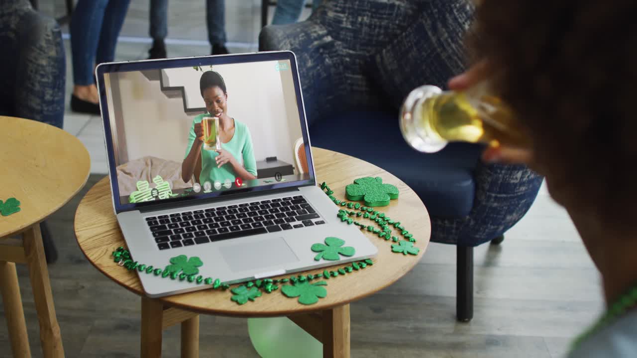 Smilende afroamerikansk kvinde med øl, iført kløverformet bånd, på videoopkald via laptop.