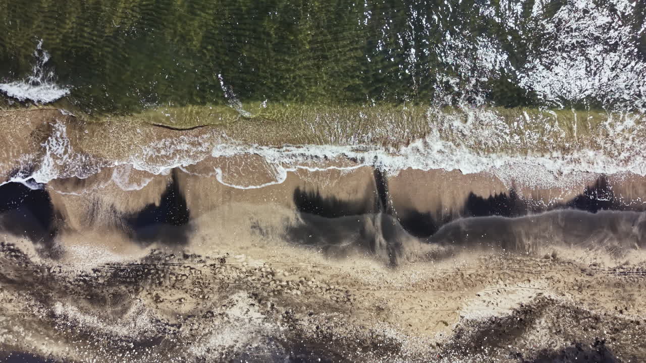 Waves crash at the shoreline with sand patterns and water reflections during daylight hours