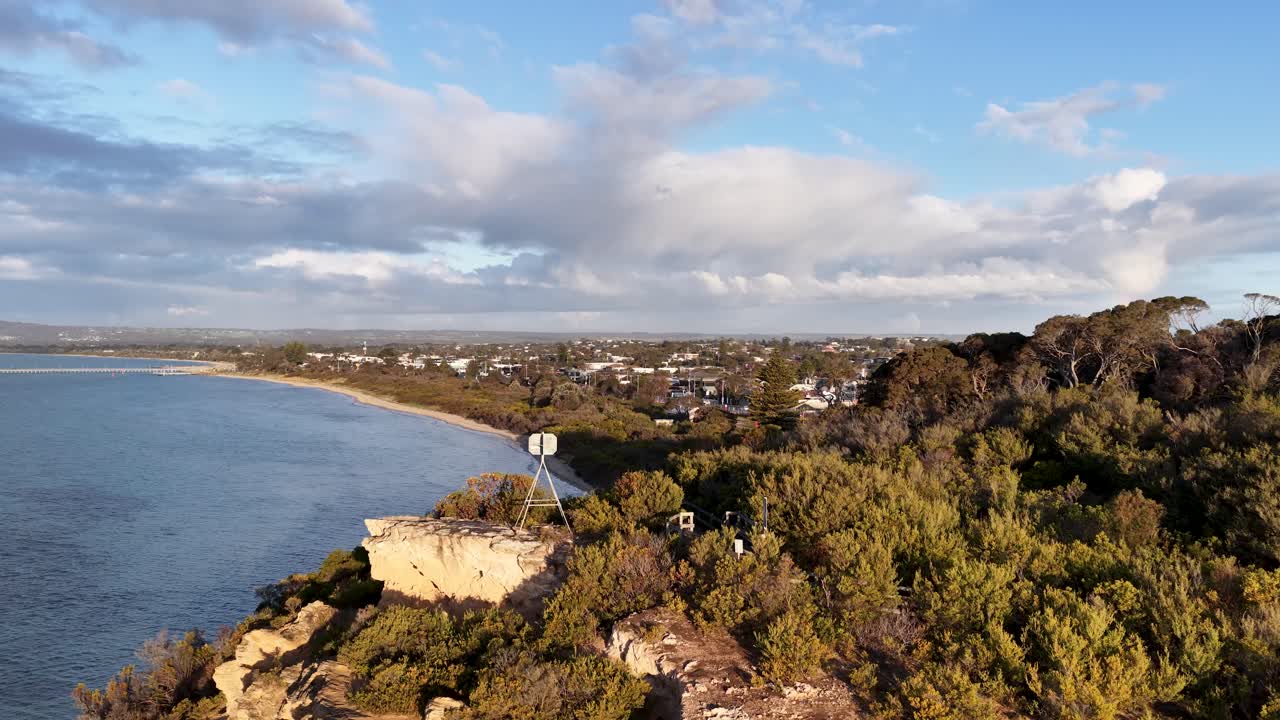 Drone glides above coastal cliffs, sandy shoreline, and residential area in golden afternoon sunlight