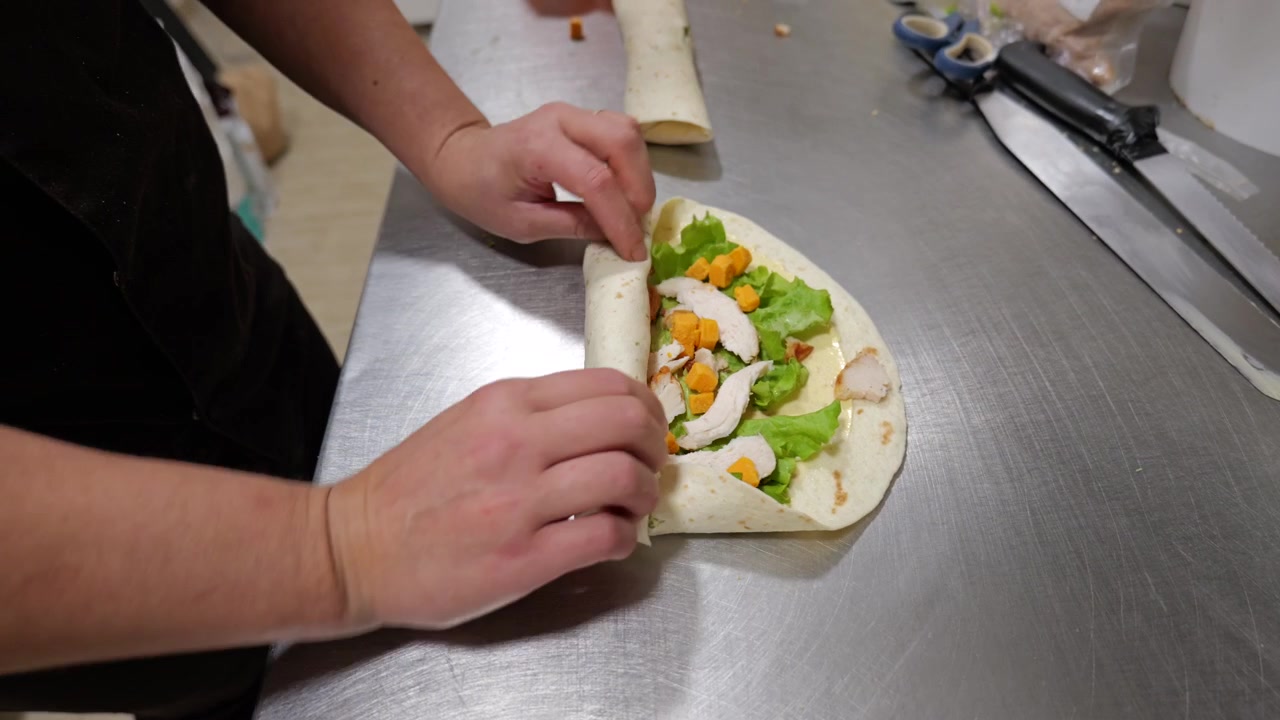 Chef assembles and rolls chicken wrap with lettuce and cheese on steel counter.