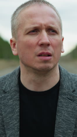 a close view of an aged white man looking focus, wearing a grey suit and a black polo, walking on a railway with stones, the background includes a blur of trees and electric poles on either side