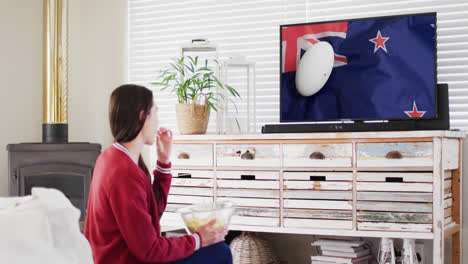 donna caucasica che guarda la tv con una palla da rugby sulla bandiera della nuova zelanda sullo schermo