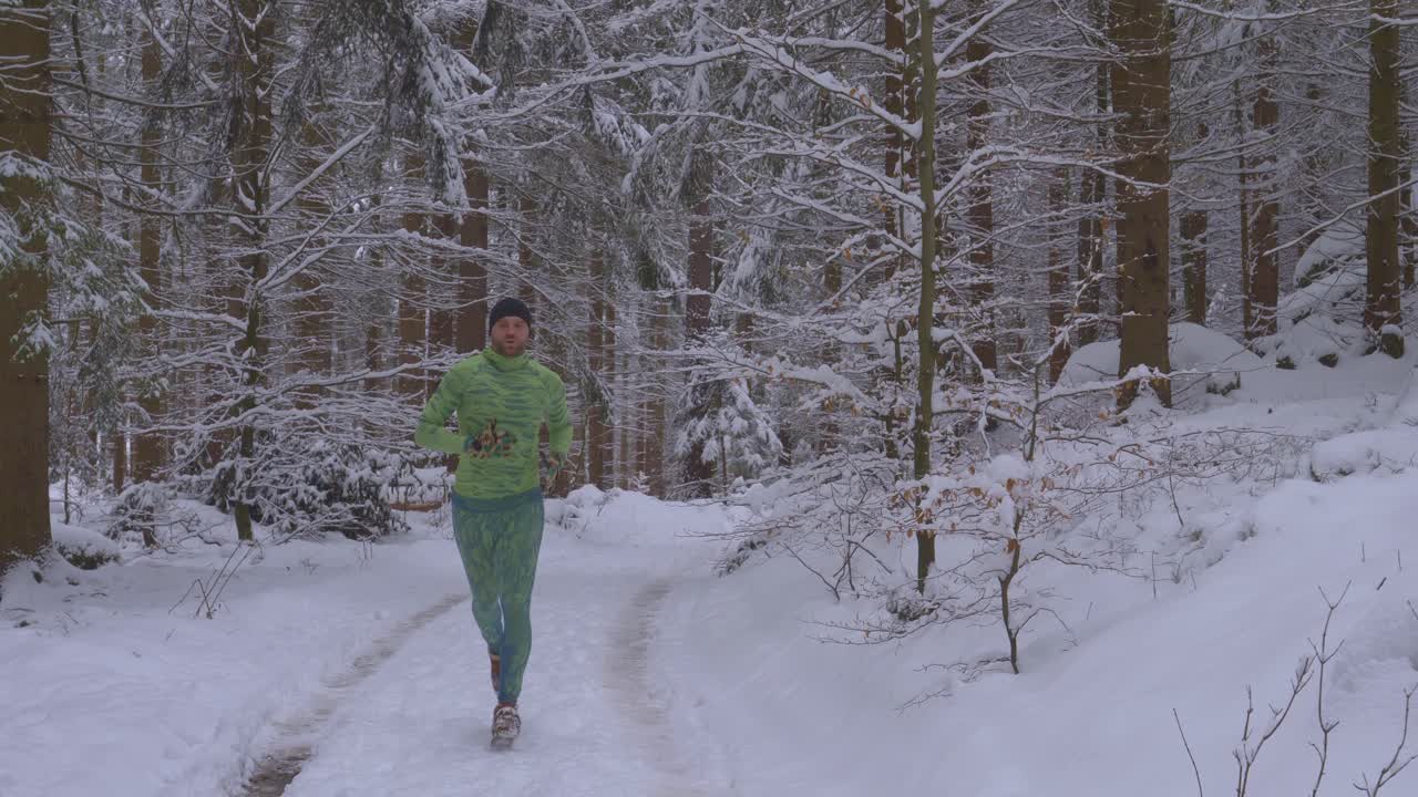 Homem barbudo correndo com roupa verde neon em floresta nevada em trilha selvagem durante um dia frio de inverno - plano de acompanhamento, em direção à câmera, câmera lenta.