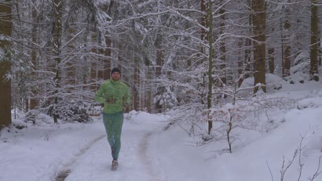 Beared-guy-running-in-neon-green-outfit-in-snowy-forest-on-wild-path-during-cold,-winter-day---follow-shot,-toward-camera,-slow-motion