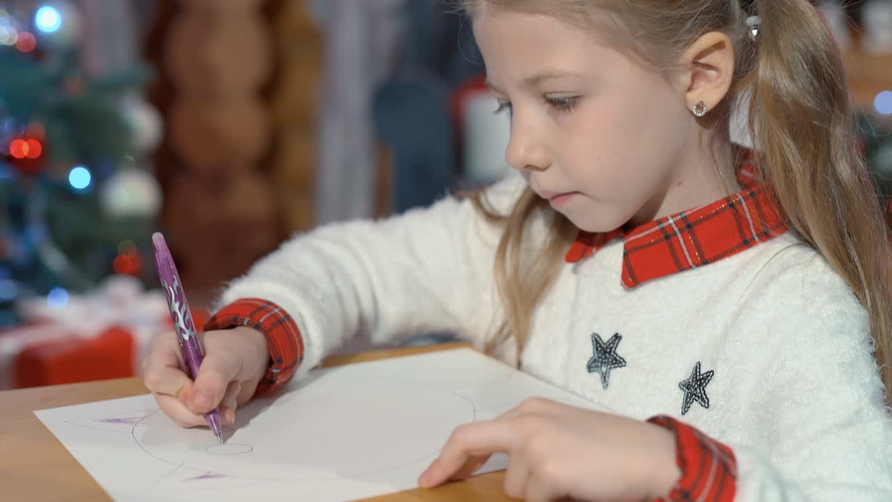 Cute little girl draws a kitten with a pencil on paper at home. Portrait of a child in a room on the background of a Christmas tree