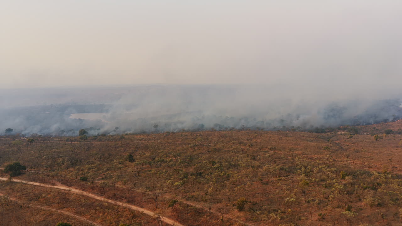 Una vasta savana arida si estende in lontananza mentre il fumo si disperde sul paesaggio, suggerendo un incendio controllato o un incendio selvaggio
