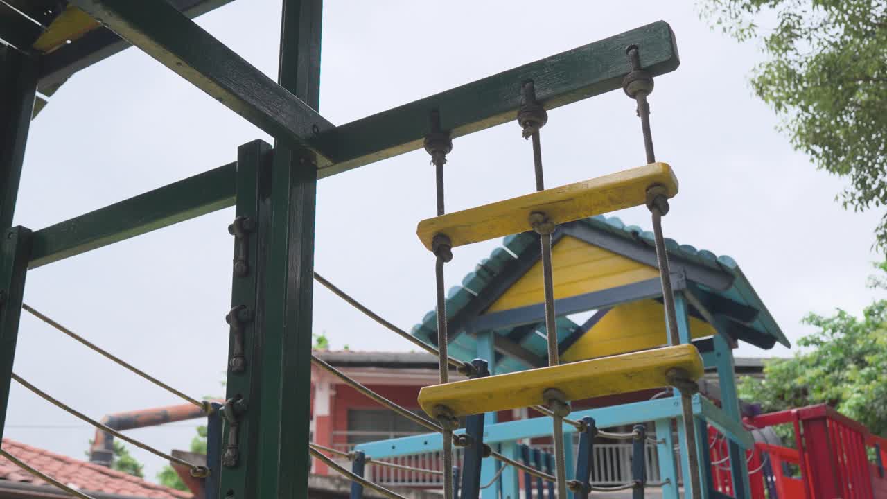 Close-up of colorful climbing structure at children's playground. Green metal frame supports yellow rope ladder. Background shows blue house with red roof and green trees. No people visible
