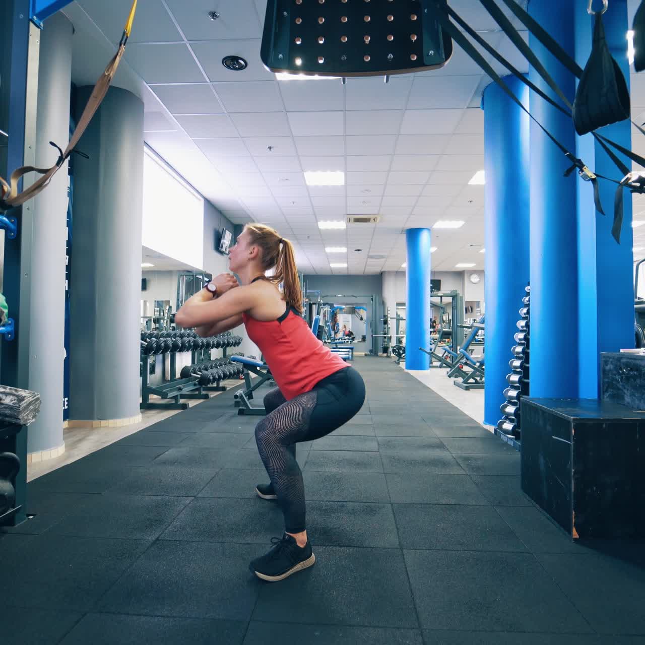 Young woman is squatting in the gym. Attractive girl doing her daily workout on fitness centre background. Healthy lifestyle.
