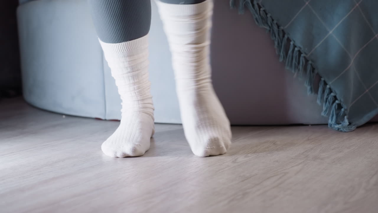 Primo piano dei piedi di un bambino con calzini bianchi che salta sul pavimento in legno vicino al letto con coperta blu, un momento accogliente in casa che simboleggia comfort, relax, atmosfera spensierata dell'infanzia e uno stile di vita casalingo semplice