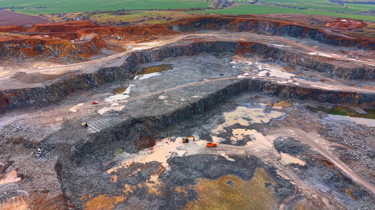 Mining site progress in rural area. Workers operate heavy machines in a large mining area with exposed rocks and puddles during daylight hours