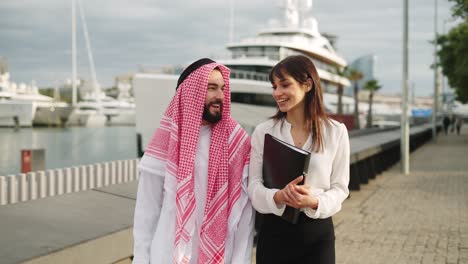 young caucasian businesswoman talking with arab man, discussing contract terms while walking in port