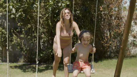 happy mother in swimsuit swinging boy on wooden swing in the backyard