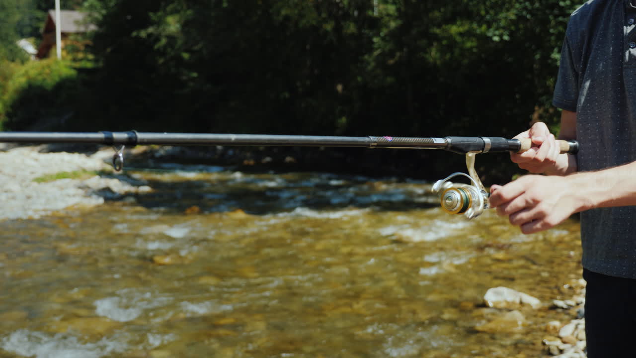 Seorang pria muda sedang memancing di sekitar sungai kecil, terlihat dari samping, hanya tangan yang tampak dalam bingkai.