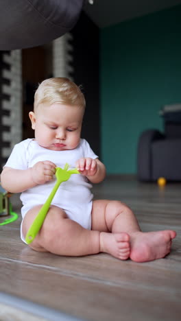 serious plump caucasian baby wearing white bodysuit sits on the floor. focused baby plays with a kitchen brush. vertical video.