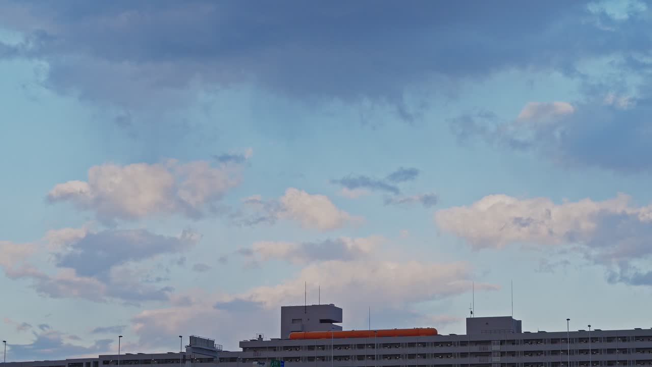 A wide-angle time-lapse showing white and grey clouds shifting quickly across a blue sky above a modern apartment complex in an urban area