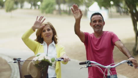 happy senior couple sitting on bikes and waving hands at camera