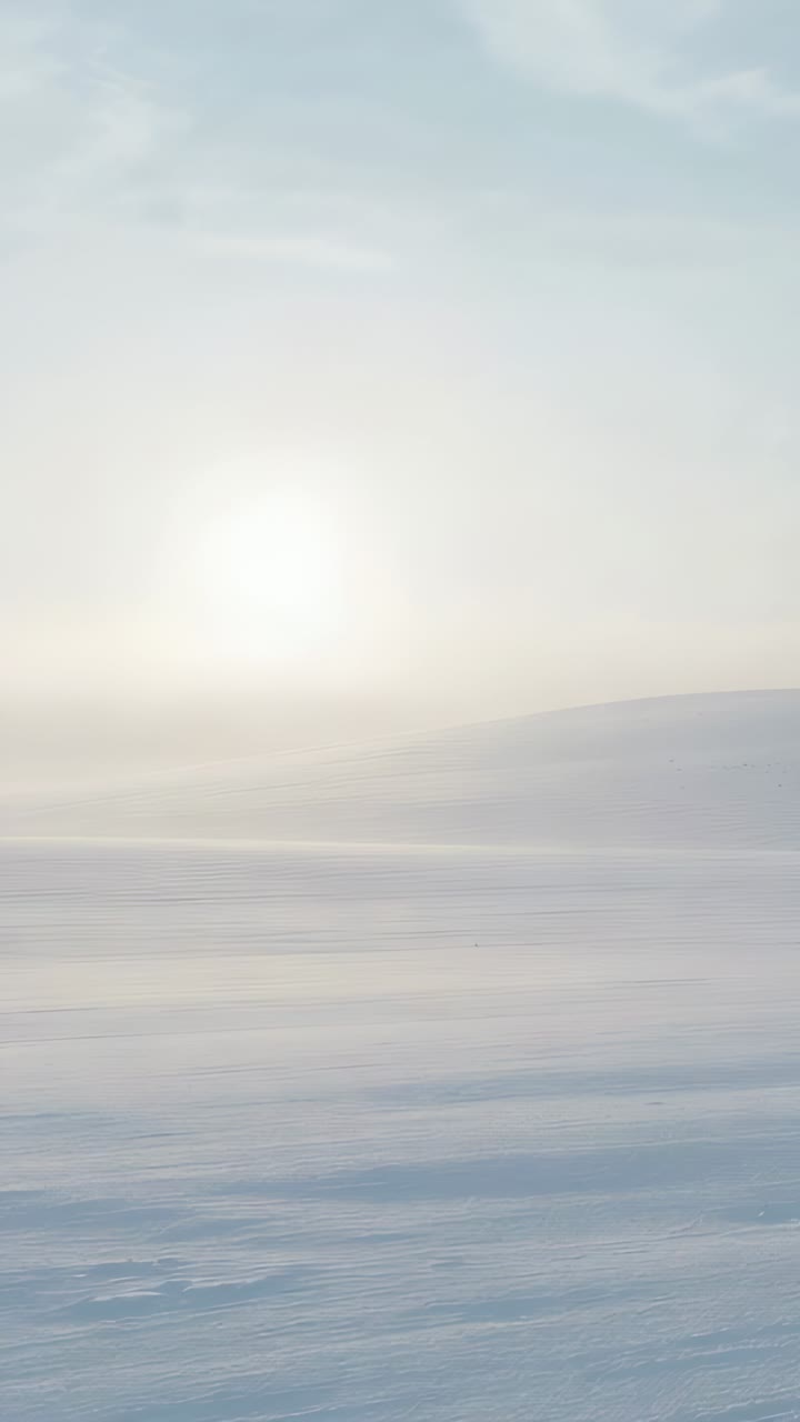 Vertical video: Rising sun casting pale glow across snow plain, revealing haze and distant ridge