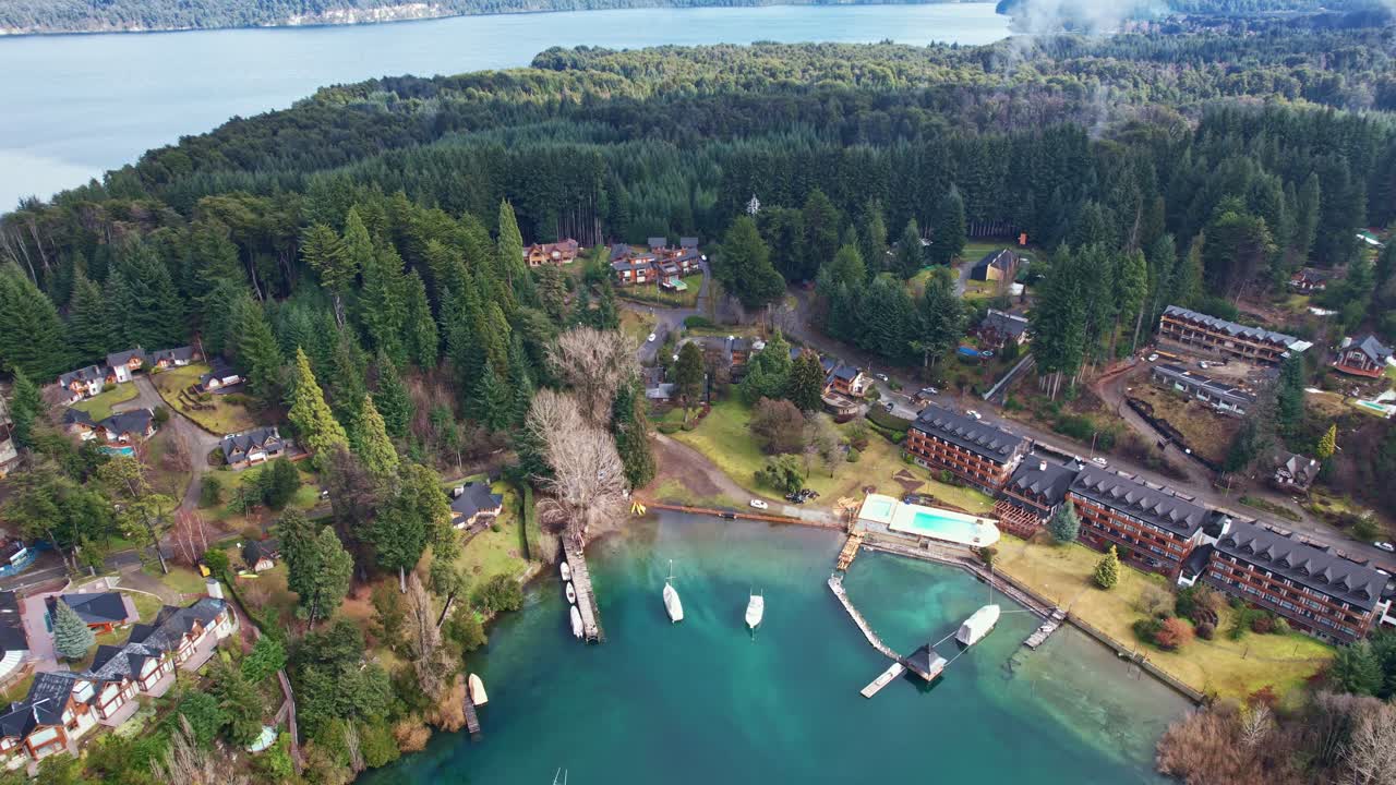 Drone che avanza su un resort di lusso sul lago e una baia. L'acqua trasparente color turchese rivela il fondale del lago, circondato da lussureggianti pinete e dalle Ande in Argentina.