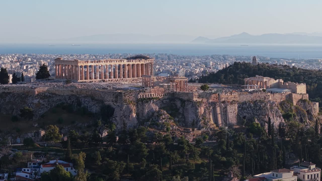 Drone shot captures the Acropolis as it rises in view, gradually revealing the cityscape of Athens and the sea stretching across the horizon in the background