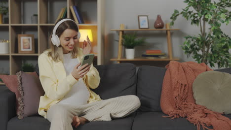 pregnant woman video calling with headphones on a couch