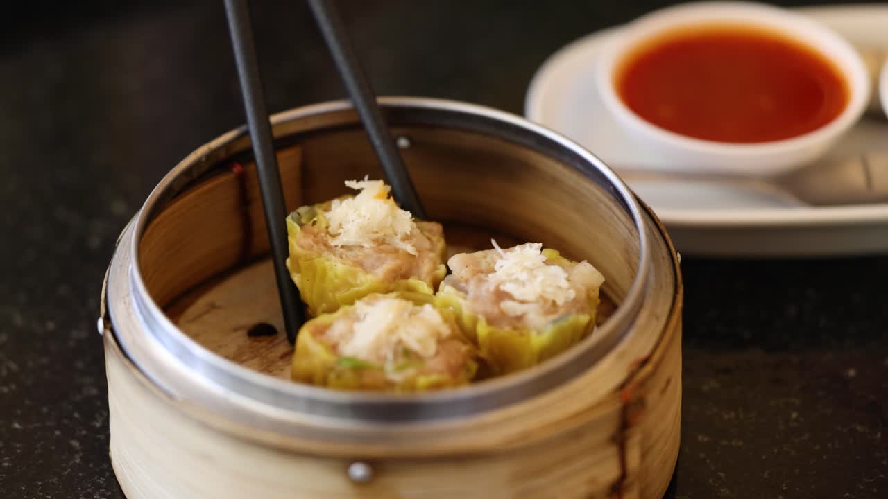 Sumpit dengan anggun mengambil dim sum dari kukusan bambu, dengan hidangan saus berwarna cerah di latar belakang, menampilkan seni kuliner Asia.