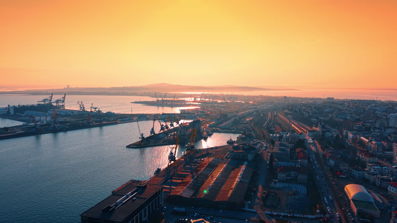 Aerial view of industrial sea port at vibrant orange sunset. Top view of cargo terminal, shipping cranes, and docks during golden hour. Calm water and hazy city horizon
