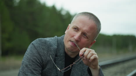 a close-up view of a middle-aged man adjusting his glasses while looking thoughtfully into the distance, he is outdoors, wearing a gray jacket, with a serene forest background