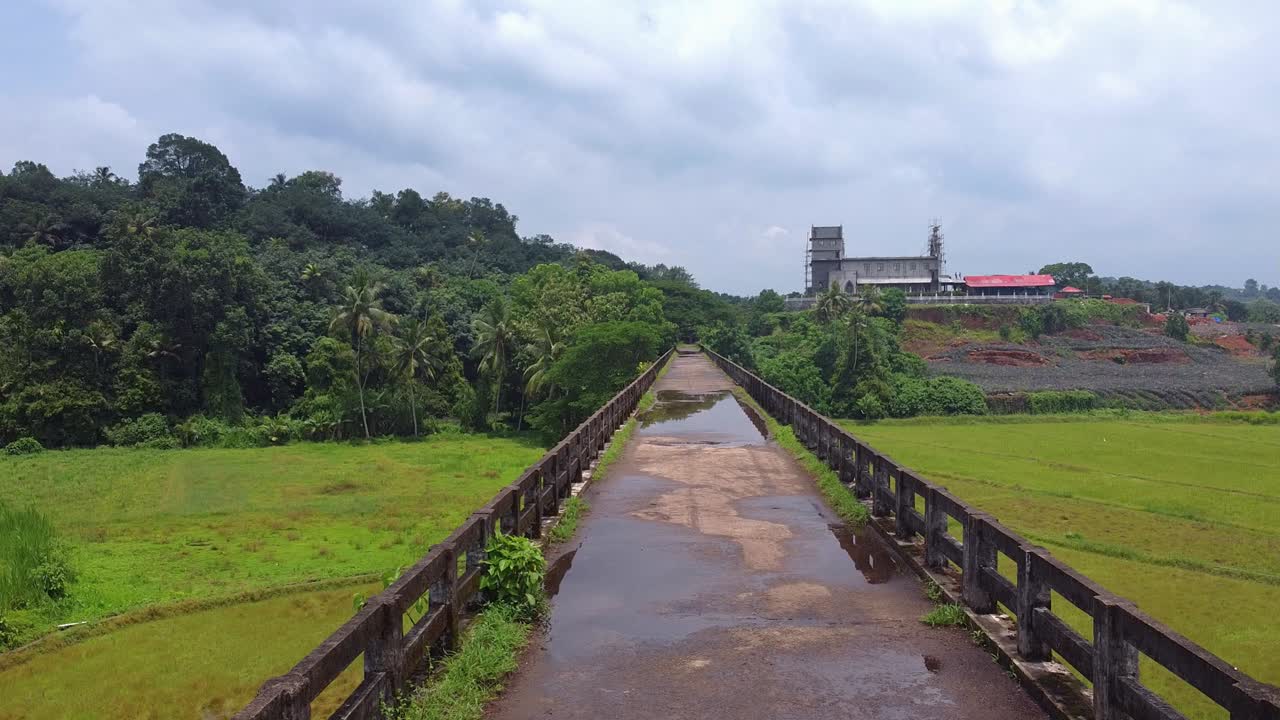 Aerial shot of a narrow, wet elevated road aqueduct over paddy fields, leading toward a large, modern Christian church under construction on a tropical hillside in Kerala