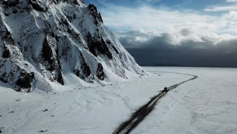 Vista-Aérea-Por-Drones-De-Un-Camión-Conduciendo-En-Un-Paisaje-Blanco-Nevado-Junto-A-Una-Imponente-Montaña-Volcánica-En-Islandia