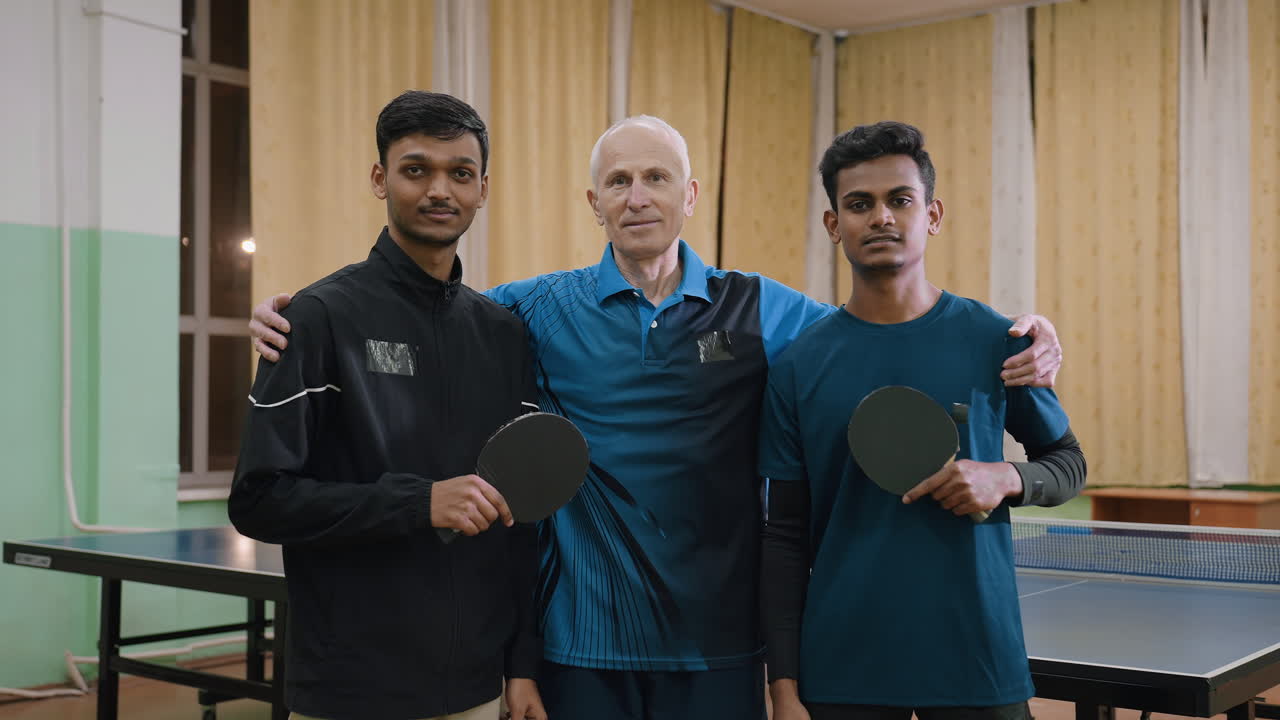 Coach places hand on young players shoulders while they pose confidently with rackets in sports hall, symbolizing mentorship, teamwork, guidance, discipline, and shared athletic achievement