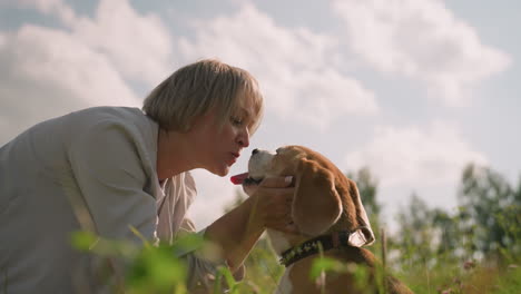 woman gently holds her dog's head and leans in to kiss the dog s open mouth in a sunny grassy field under a bright sky, capturing a tender moment of affection