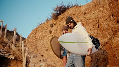 A-brunette-man-in-a-white-sweatshirt-helps-his-blonde-girlfriend-in-a-checkered-shirt-go-down-the-stairs-near-the-rocky-seashore.-They-carry-surfboards-in-their-hands-and-walk-to-the-sea-in-the-morning-at-Sunrise