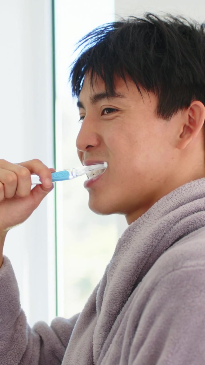 Vertical video: Smiling man in gray robe lifting blue brush, brushing teeth in bathroom for morning