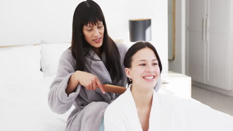 happy biracial mother in bathrobe brushing hair of smiling adult daughter in sunny room, slow motion