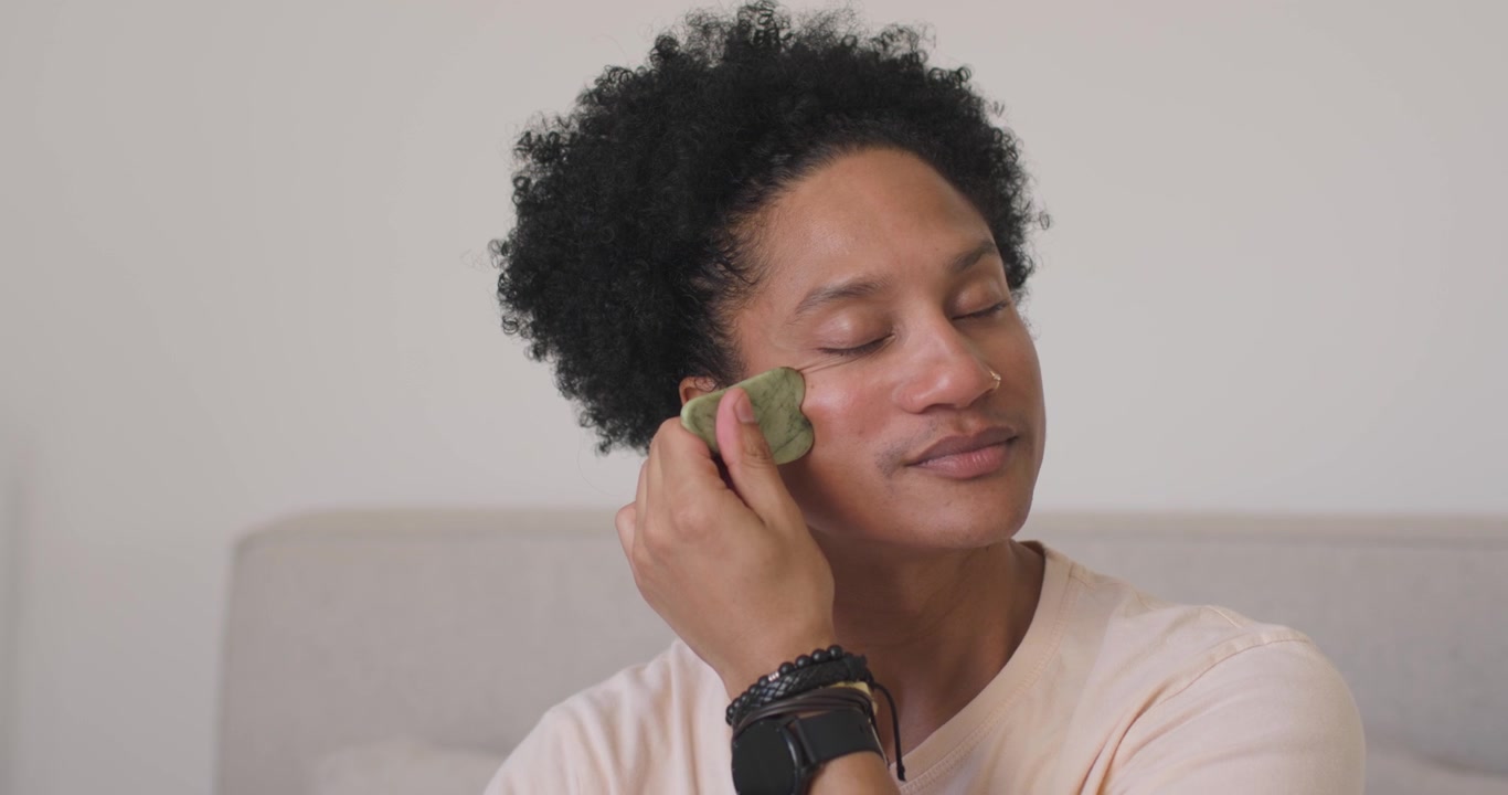 African American adult man glancing at mirror, massaging cheek with gua sha on sofa for skincare