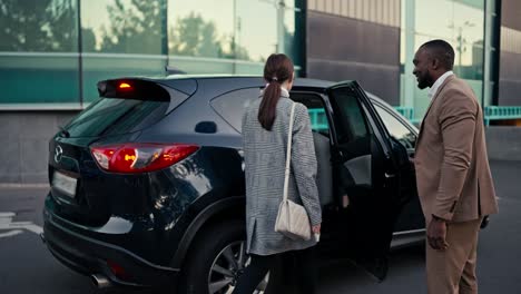 Confident-and-happy-man-Businessman-with-Black-skin-and-a-beard-in-a-brown-suit-invites-a-brunette-female-colleague-to-sit-in-the-passenger-seat-of-a-black-car-and-sits-behind-her-during-their-business-trip-in-the-city