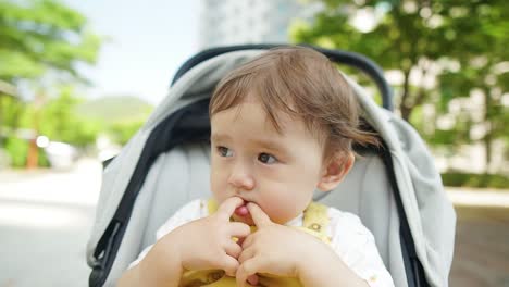 Adorable-Niña-De-Etnia-Mixta-Chupándose-Los-Dedos-Mientras-Se-Sienta-En-Un-Cochecito-Al-Aire-Libre