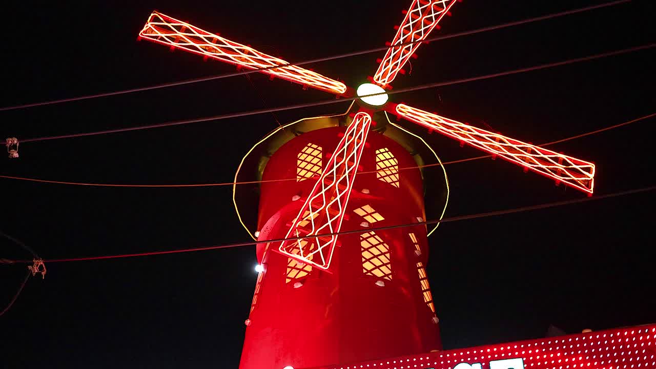 A vibrant red windmill with glowing blades spins against a dark night sky, creating a dynamic and colorful scene