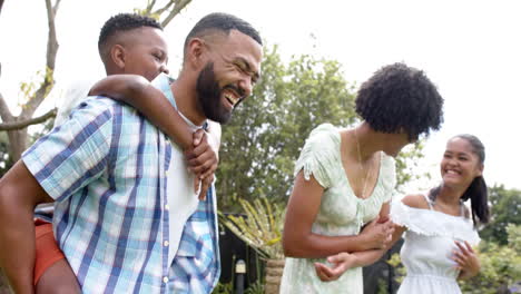 Happy-african-american-parents,-daughter-and-son-piggybacking-and-walking-in-garden,-slow-motion