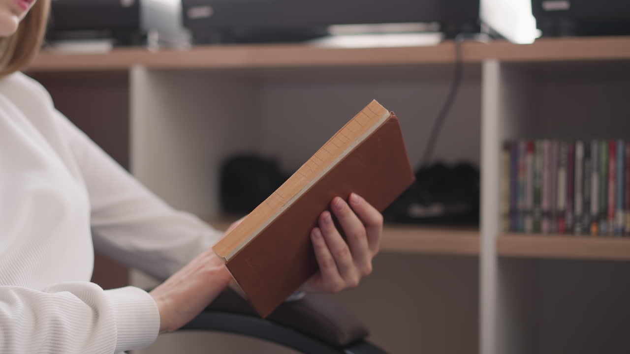 libro en manos de una joven lectora de primer plano. mujer se sumerge en la lectura girando pensativamente la página del libro en el almacenamiento de libros circundante. mujer con novela iluminada