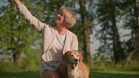 woman taking selfie with her dog in grassy field under warm sunlight, dog focused on something else while owner smiles happily, surrounded by trees and greenery