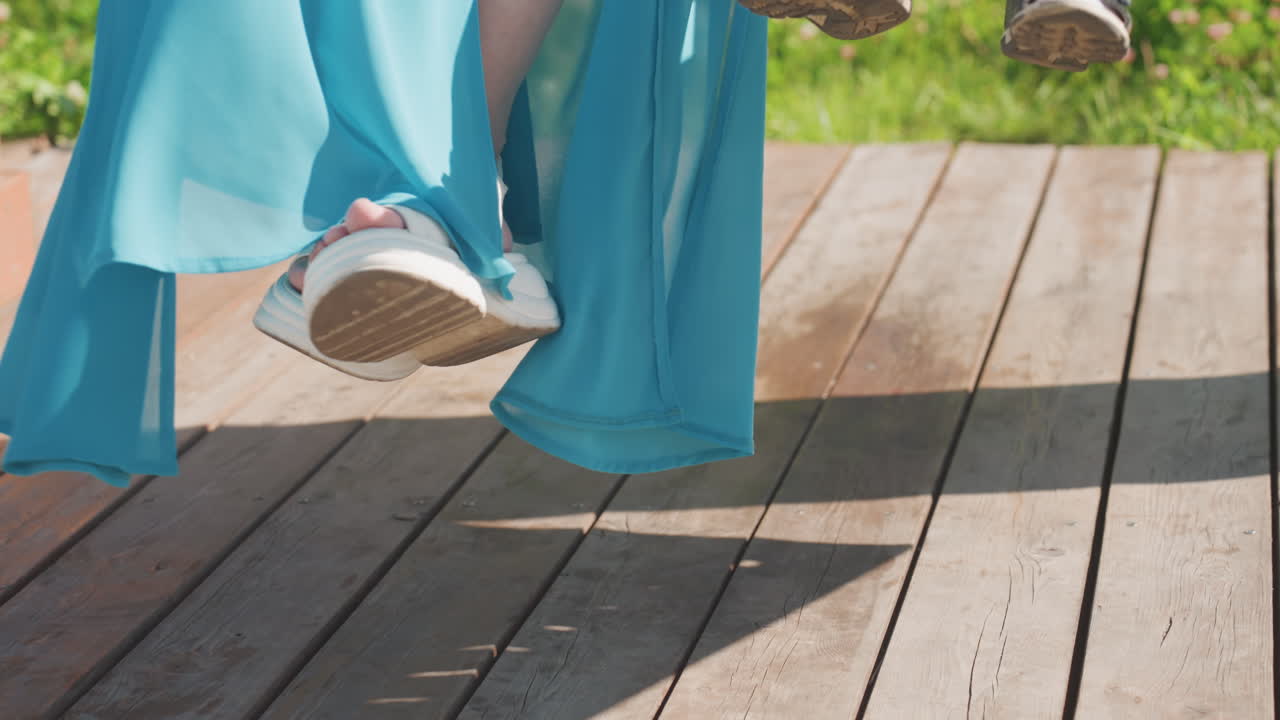 Kvinde svinger fødderne over træterrasse, flydende blå kjole og hvide sandaler fanger sollys. Barnefødder er synlige ved siden af den voksne, blød skygge på brædderne. Et afslappet familie weekendøjeblik.