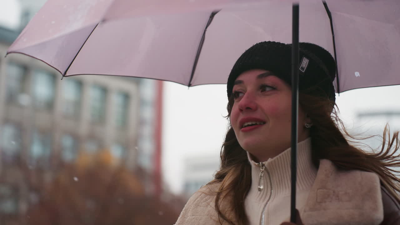 Upper body shot of student smiling and walking in light snowfall holding umbrella wearing knit cap brown shearling jacket, cold weather, casual, happy, urban background, peaceful, winter
